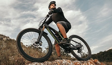 Male cyclist in dark clothing sits on his black and green electric mountain bike on top of a rockface