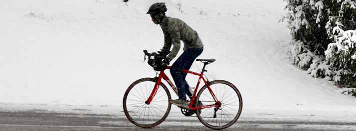 A male cyclist wearing dark clothing and a black helmet rides a red bike in the middle of winter